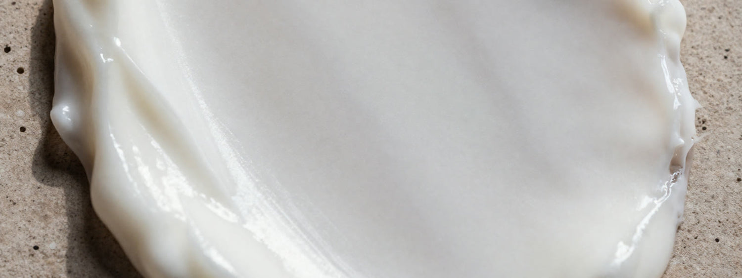 Macro de um disco de creme branco espalhado sobre laje de pedra clara e porosa, com a borda ondulada do creme atravessando o quadro.