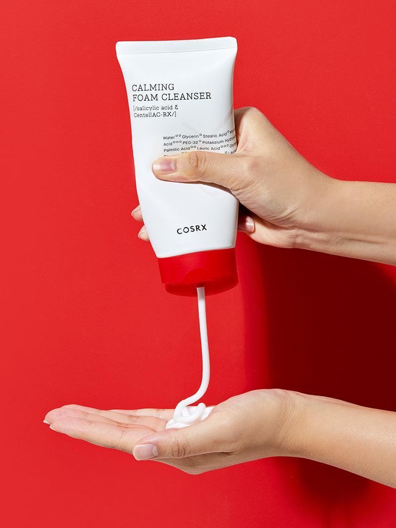 Maos segurando a bisnaga e dispensando o produto na palma, sobre fundo vermelho