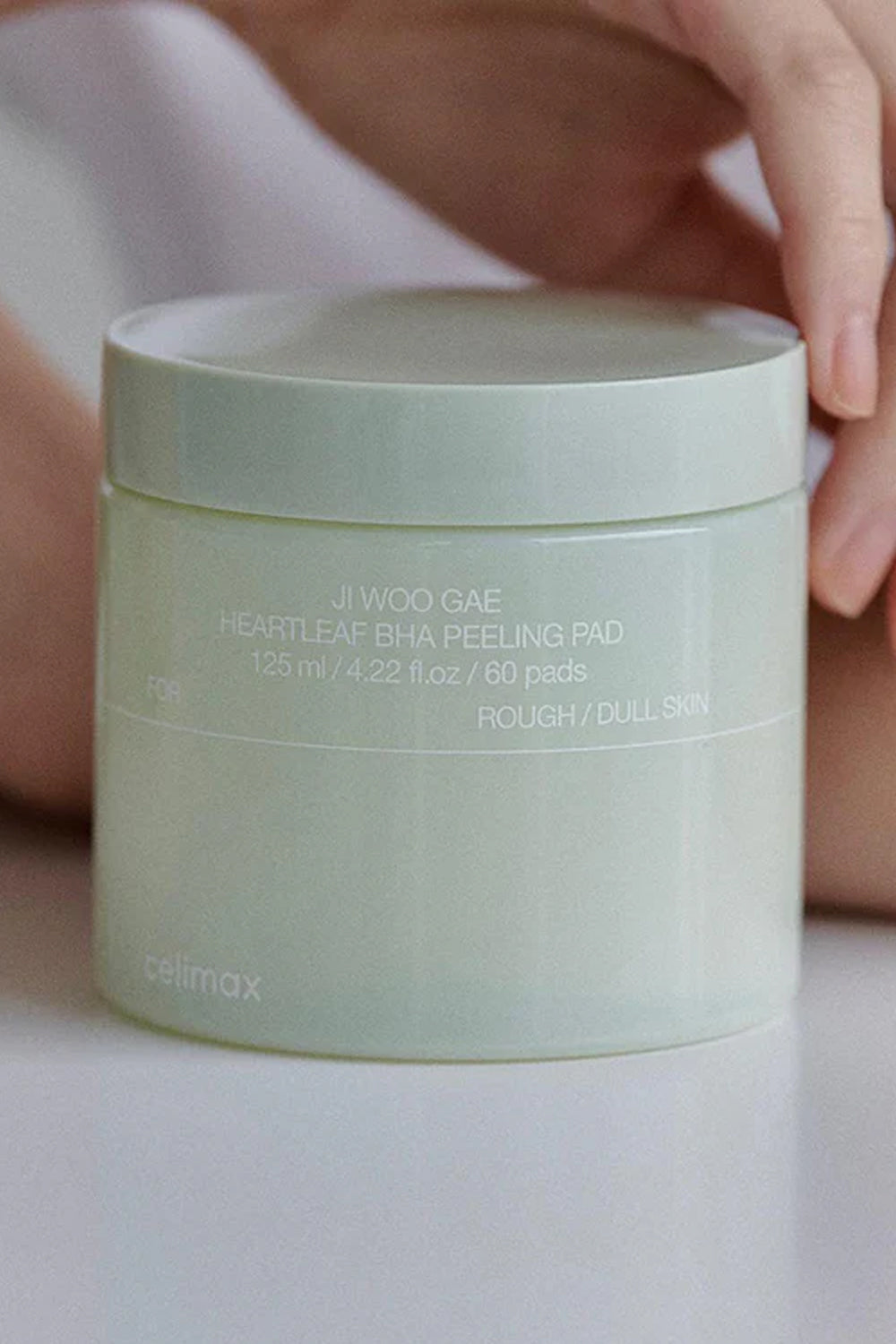 Mao de modelo apoiada atras do pote verde do Celimax Ji Woo Gae Heartleaf BHA Peeling Pad.