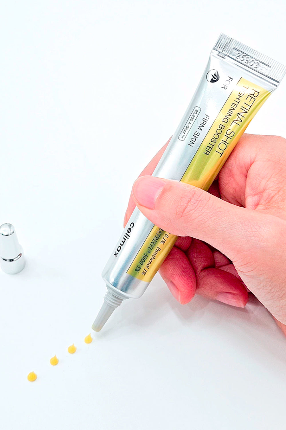 Mao segurando a bisnaga do Celimax The Vita-A Retinal Shot, com gotas amarelas do produto sobre a superficie branca.