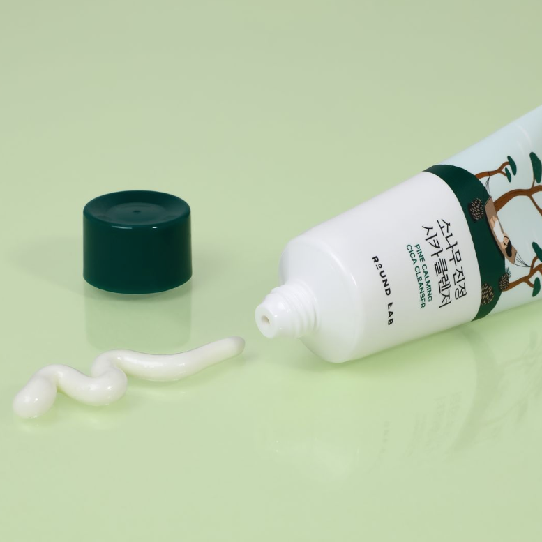 Bisnaga branca do cleanser Pine Calming Cica deitada ao lado de um filete do produto, fundo verde claro.