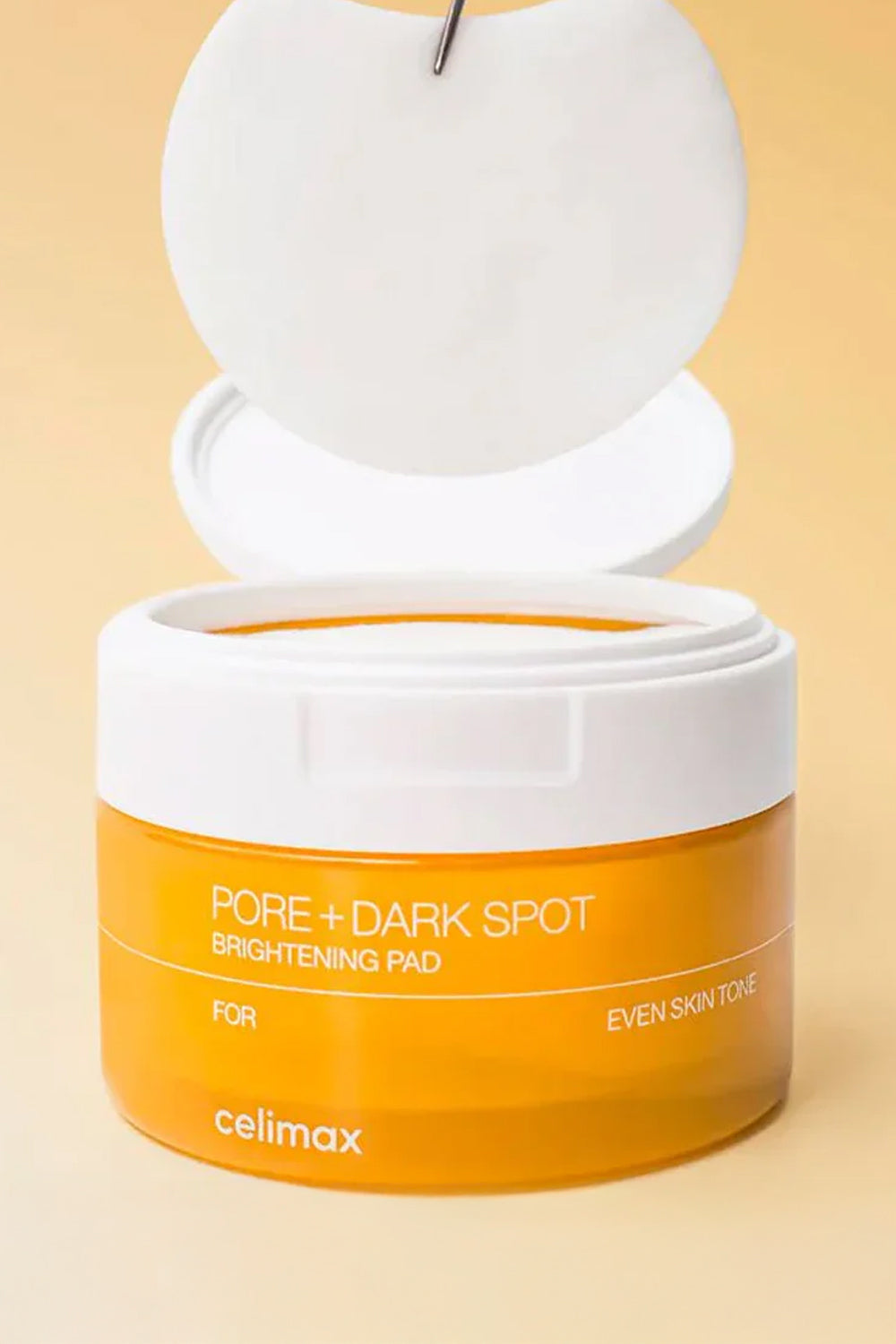 Pote Celimax Pore + Dark Spot aberto com um disco de algodão suspenso acima, sobre fundo bege