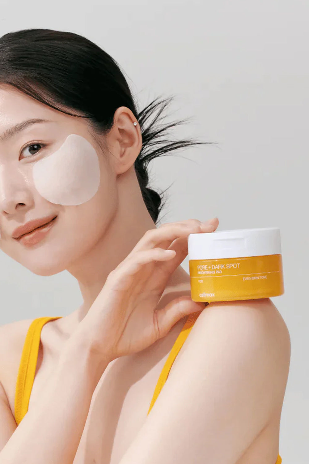 Modelo adulta com um disco de algodão no rosto, segurando o pote Celimax Pore + Dark Spot
